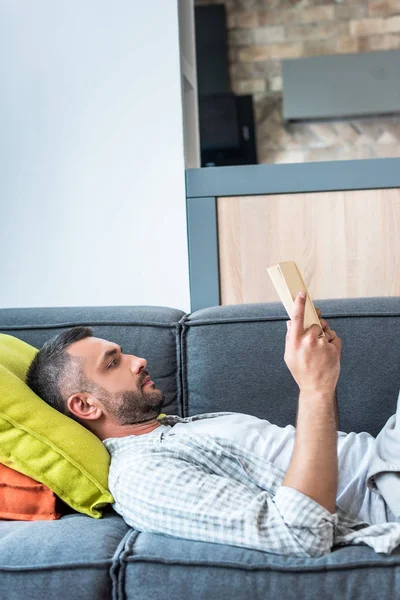 Vista Lateral Homem Barbudo Livro Leitura Enquanto Deitado Sofá Casa — Fotografia de Stock Grátis