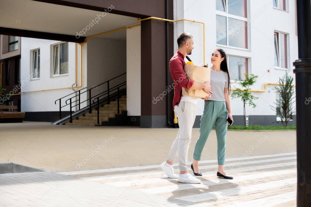 man with paper package full of vegetables and woman with smartphone in hand crossing road together on street