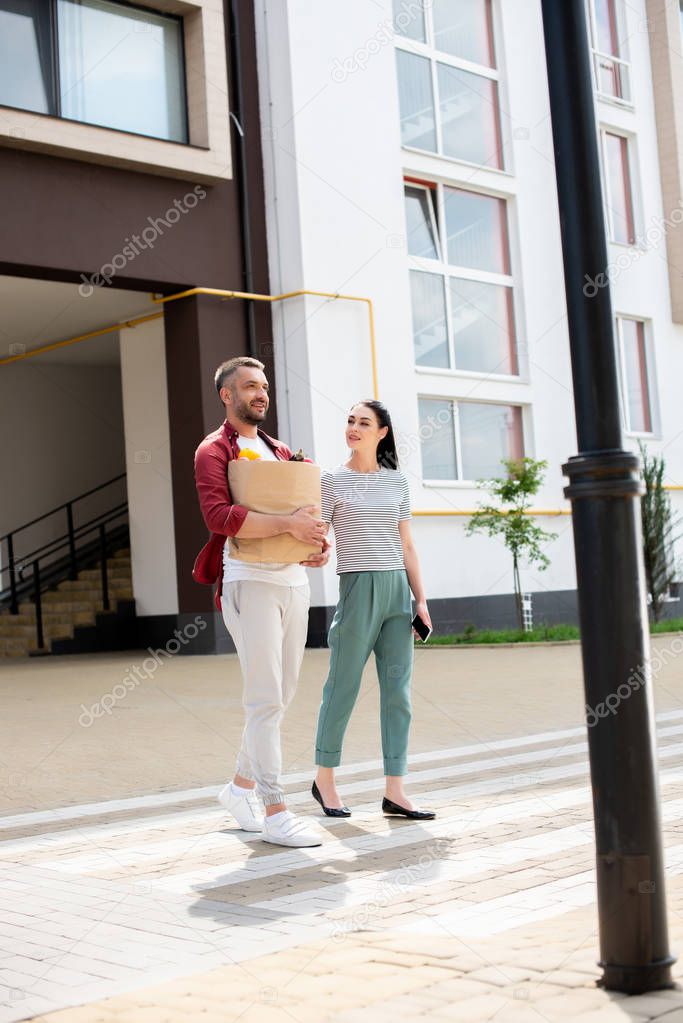 man with paper package full of vegetables and woman with smartphone in hand crossing road together on street