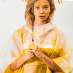 Atractiva mujer en impermeable pintado con pintura amarilla de pie bajo paraguas aislado en blanco