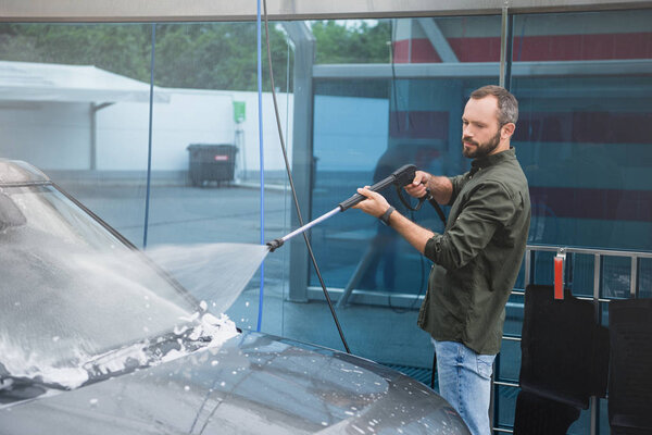 handsome man cleaning car front window at car wash with high pressure water jet