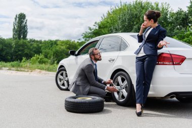 handsome businessman changing tires on car on road, businesswoman talking by smartphone clipart