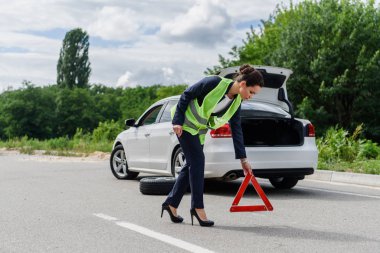 attractive businesswoman in light green vest putting stop sign on road near broken car clipart
