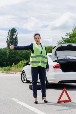 attractive businesswoman in light green vest hitchhiking on road near broken car clipart