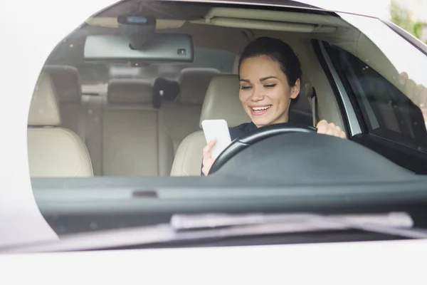 笑顔の魅力的な女性が車を運転中にスマート フォンを使用して — ストック写真