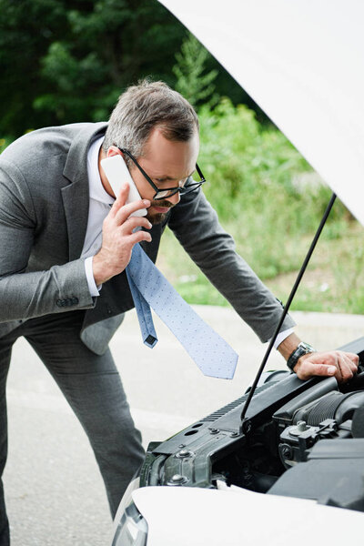 businessman talking by smartphone and looking in open car hood