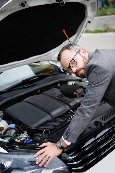 handsome businessman leaning on open car hood and looking at camera 
