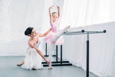 adorable child in pink tutu stretching and looking at camera while practicing ballet with teacher clipart