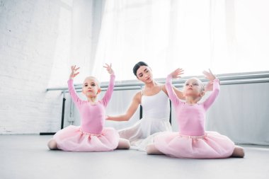 young ballet teacher looking at cute little kids in tutu skirts practicing ballet in school clipart