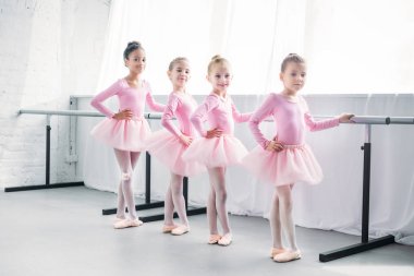 cute multiethnic kids in pink tutu skirts exercising and smiling at camera in ballet studio clipart