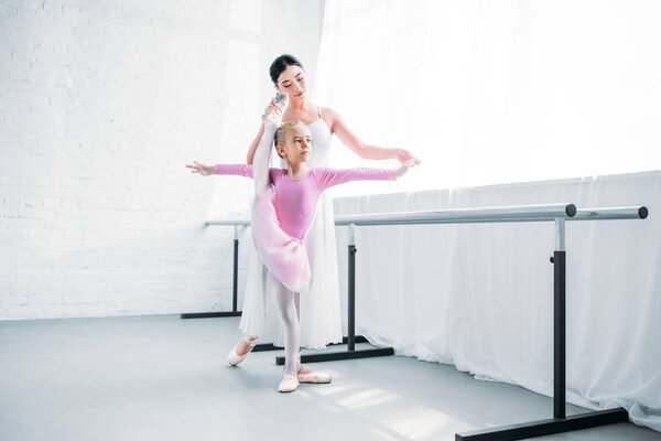 adorable little ballerina in pink tutu exercising with teacher in ballet school