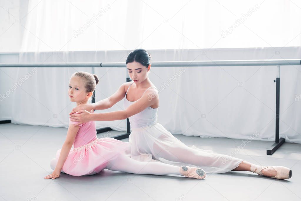 adorable little ballerina in pink tutu looking at camera while exercising with teacher in ballet school