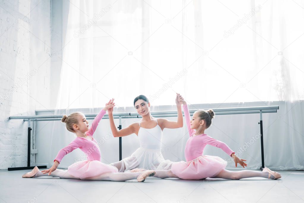 young ballet teacher with children in tutu skirts sitting and holding hands while exercising in ballet school