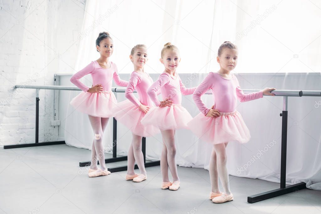 cute multiethnic kids in pink tutu skirts exercising and smiling at camera in ballet studio