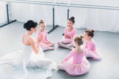 high angle view of kids in pink tutu skirts and young ballet teacher sitting together in ballet school