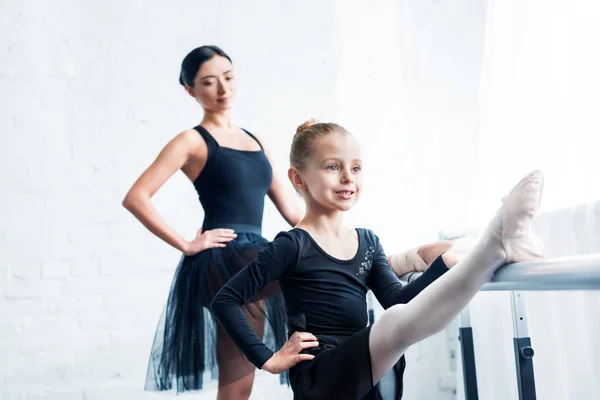 Sonriente Pequeña Bailarina Estirándose Mientras Profesora Pie Detrás Escuela Ballet —  Fotos de Stock