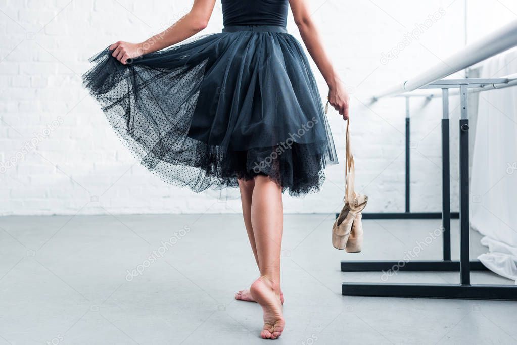 cropped shot of ballerina in black tutu holding pointe shoes in ballet studio 