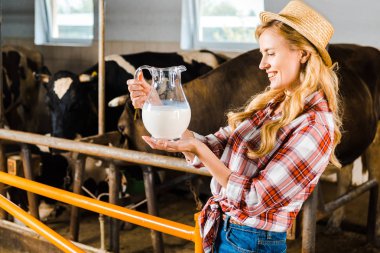 side view of attractive farmer looking at jug of milk in stable clipart