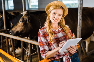 attractive farmer in straw hat holding tablet and looking at camera in stable clipart