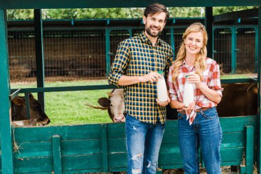 smiling couple of farmers holding bottles of cow milk near stable clipart