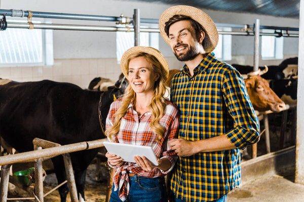 smiling couple of farmers with tablet looking away in stable