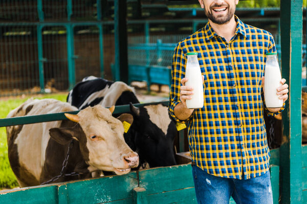 cropped image of smiling farmer holding bottles of cow milk near stable