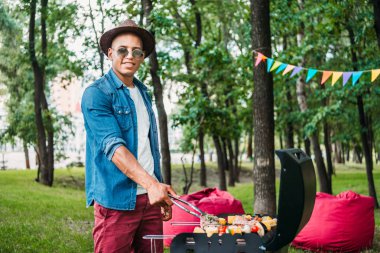Afro-Amerikan erkek güneş gözlüğü ve şapka yemek yemek ızgara Park yan görünüm