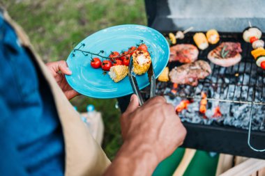 partial view of african american man wit tongs putting grilled vegetables on plate during bbq in park clipart