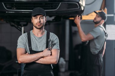 auto mechanic posing in overalls with crossed arms, while coworker working in workshop behind clipart