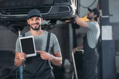 smiling mechanic showing digital tablet with blank screen, while colleague working in workshop behind