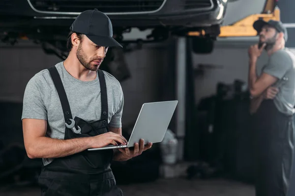 Automechaniker Mit Laptop Während Kollege Werkstatt Dahinter Arbeitet — Stockfoto