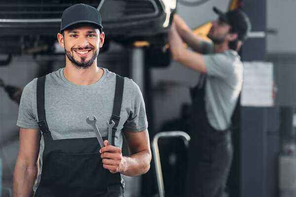 smiling auto mechanic holding wrenches, while colleague working in workshop behind