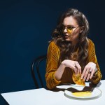 Fashionable woman with unhealthy meat pastry sitting at table with blue backdrop