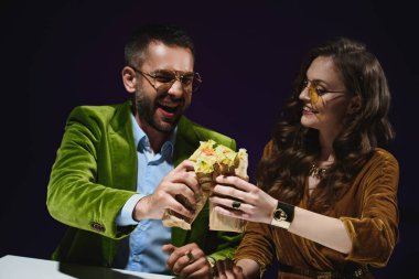 portrait of smiling couple in luxury velvet clothing with shawarma sitting at table with dark background behind  clipart