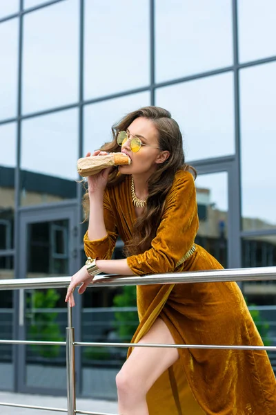 portrait of attractive woman in luxury clothing and sunglasses eating french hot dog on street
