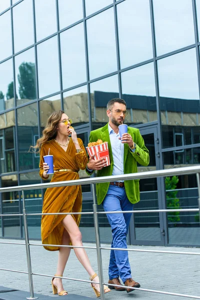 fashionable couple in velvet clothing with soda drinks eating fried chicken legs while walking on street