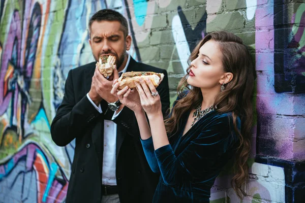 portrait of couple in luxury clothing with hot dogs standing near wall with graffiti on street