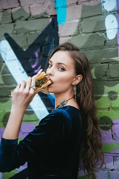 side view of beautiful woman in stylish clothing eating hot dog on street