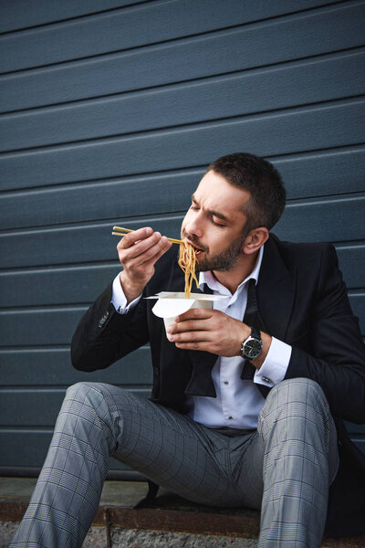 portrait of man in stylish suit with chopsticks eating asian food on street