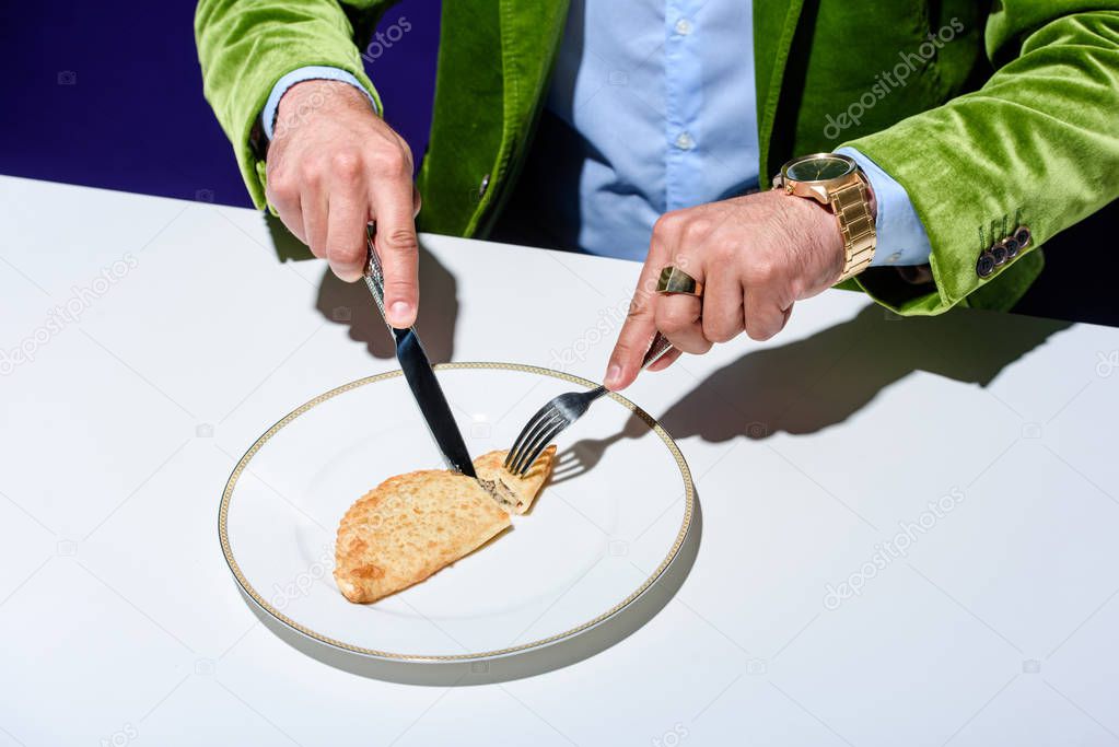 cropped shot of man in stylish green velvet jacket cutting meat pastry on plate with blue background behind