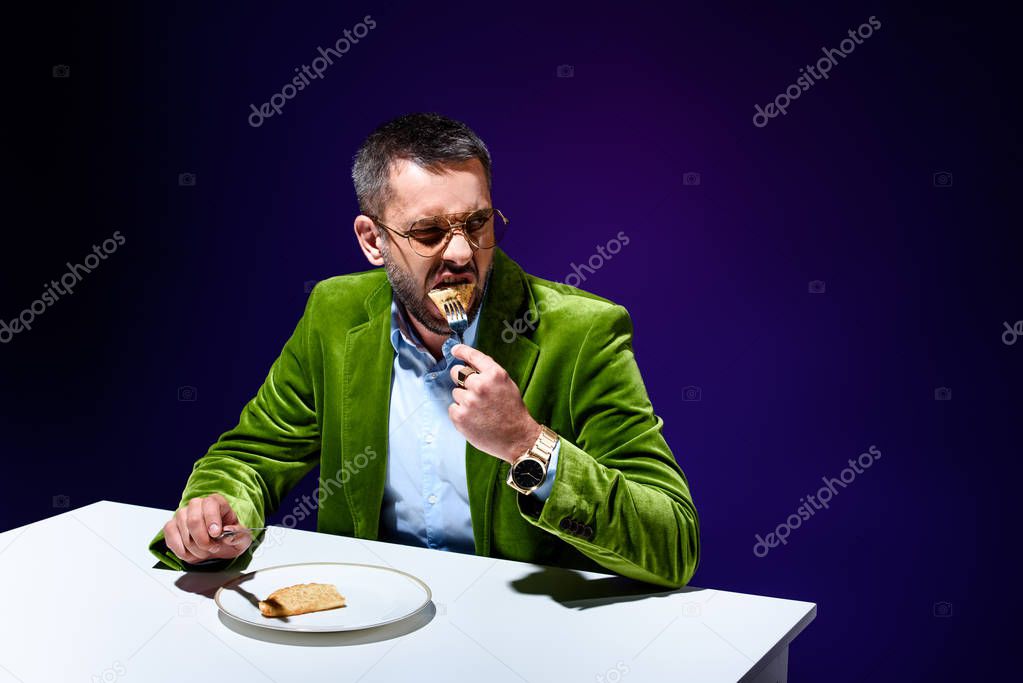 portrait of man in stylish green velvet jacket eating meat pastry on plate at table with blue background behind