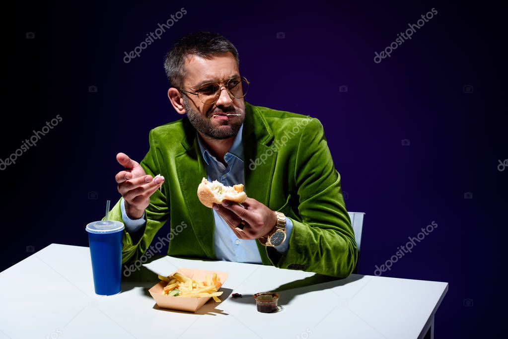 man in velvet jacket eating burger at table with french cries and soda drink with blue background