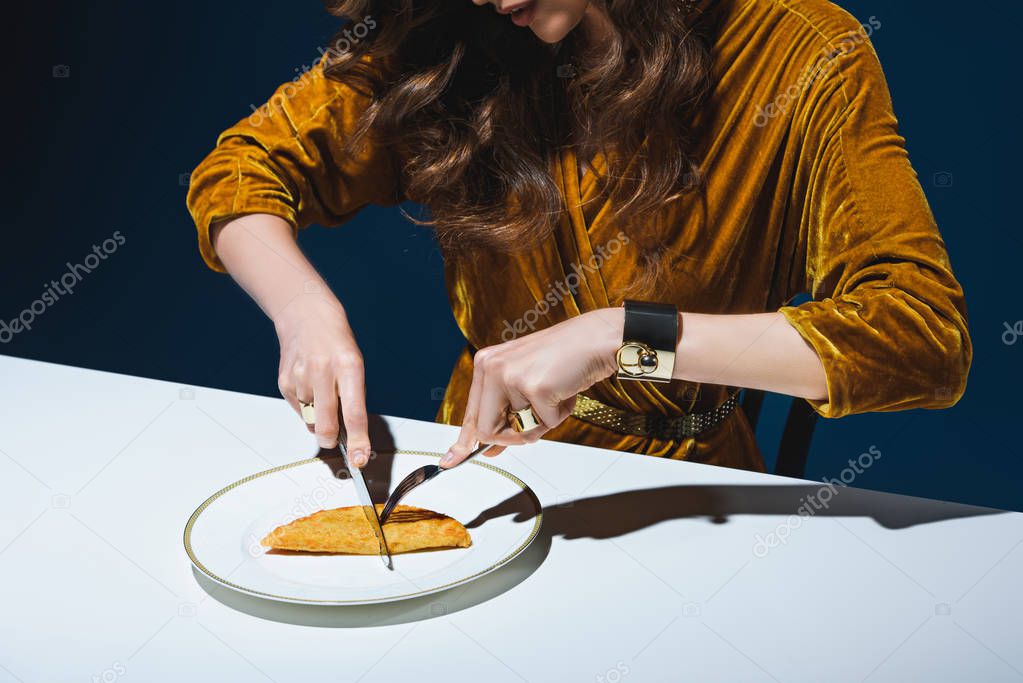 cropped shot of woman in stylish clothing cutting unhealthy meat pastry at table with blue backdrop behind