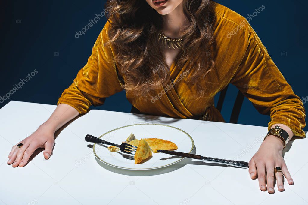 cropped shot of woman in luxury clothing sitting at table with meat pastry on plate with blue backdrop