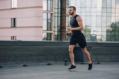 side view of handsome sportsman running and listening music with smartphone on roof clipart