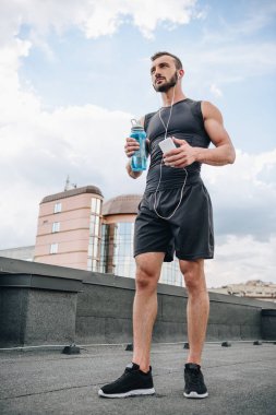 yakışıklı sporcu dinleme müzik smartphone ve spor şişe çatı üzerinde tutan düşük açılı görünüş