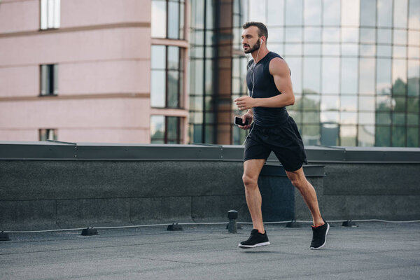 side view of handsome sportsman running and listening music with smartphone on roof