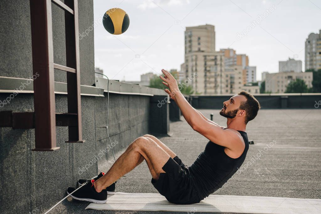 side view of handsome sportsman doing sit ups with medicine ball on yoga mat on roof