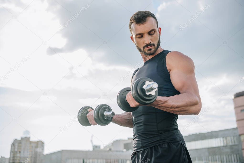 handsome sportsman training with dumbbells on roof and looking at biceps