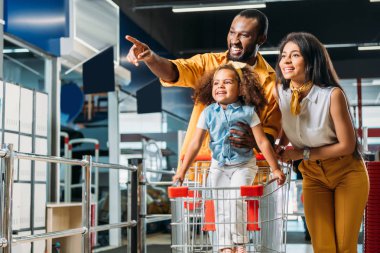 smiling african american man pointing by finger to little daughter and wife in supermarket  clipart